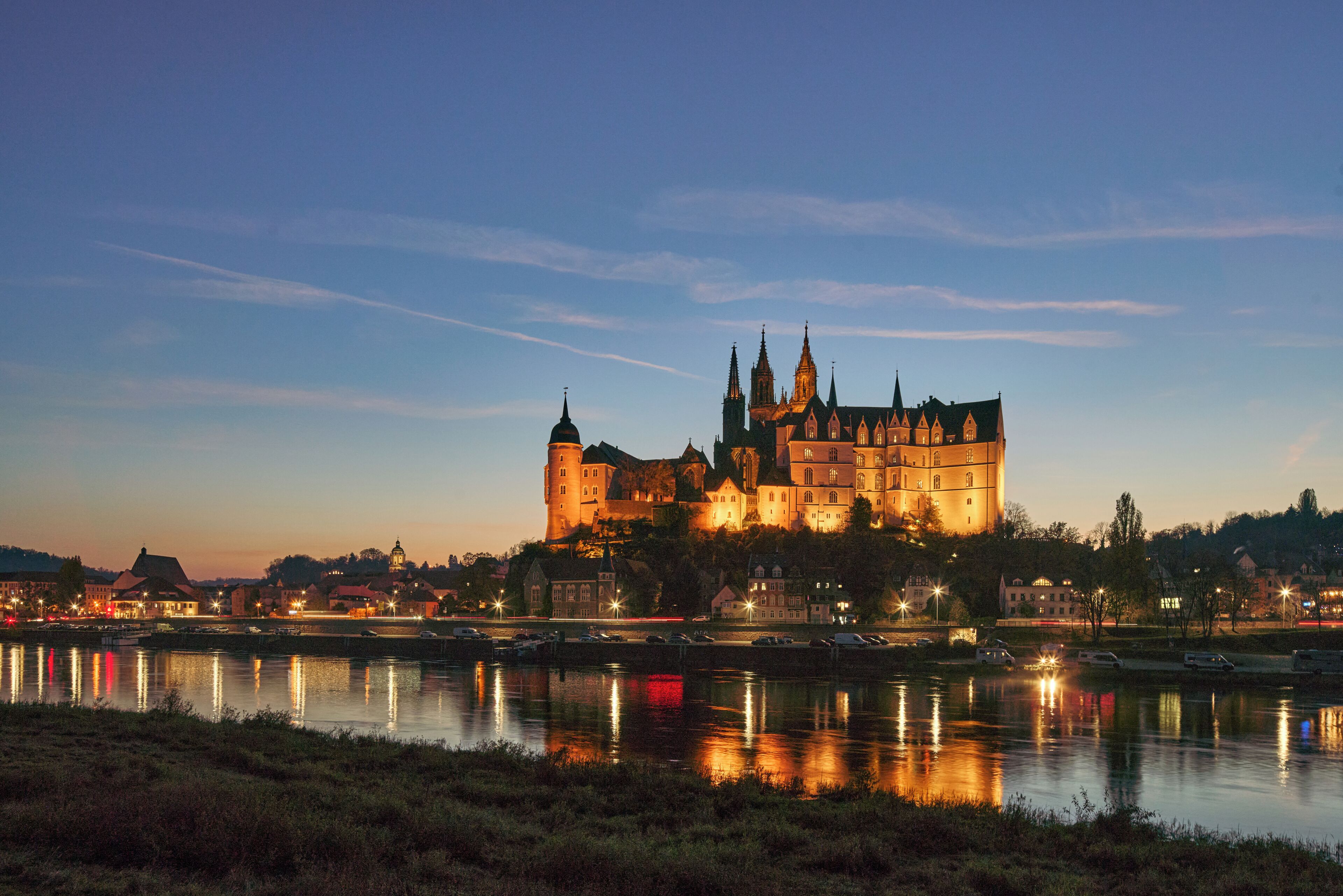 A historic castle with multiple towers, situated by a calm river reflecting city lights.