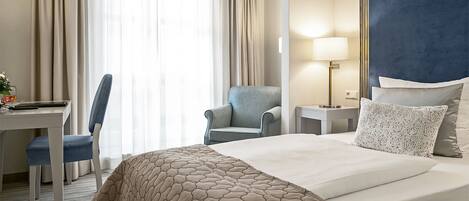 A hotel room with a bed, a desk, a chair, a lamp, and curtains.