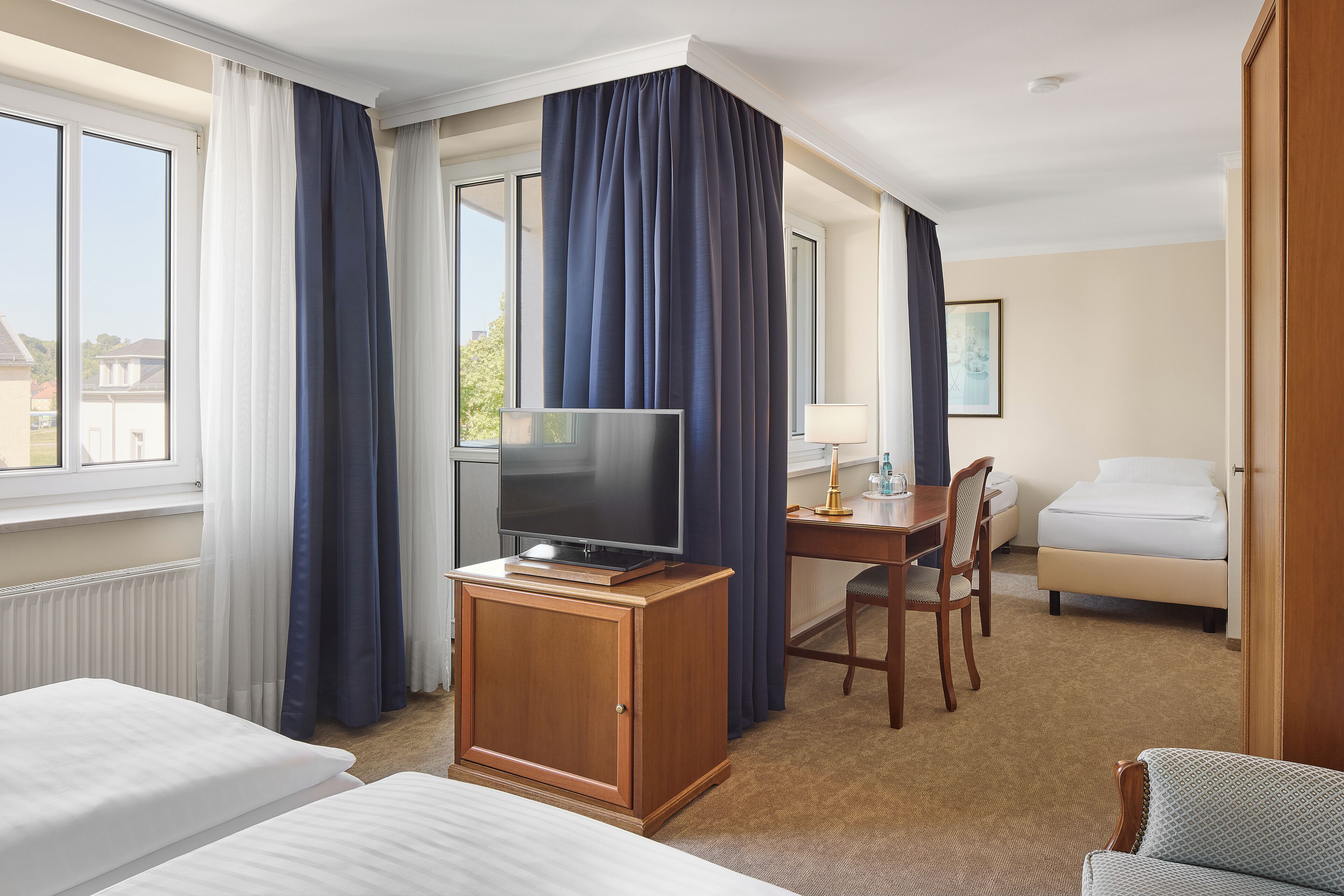 Habitación de hotel con cama, escritorio con televisor, silla, lámpara y ventana con cortinas.