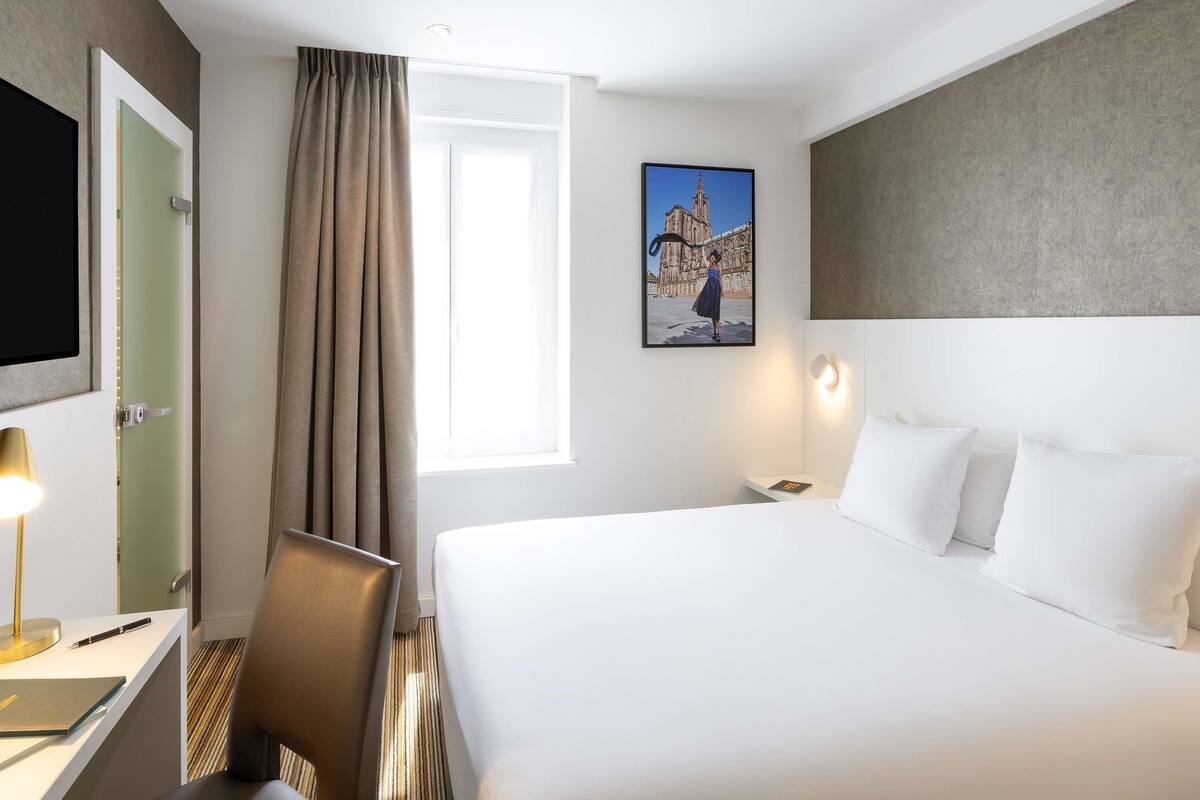 A hotel room with a bed, a desk with a lamp, a chair, a painting, and a window with curtains.