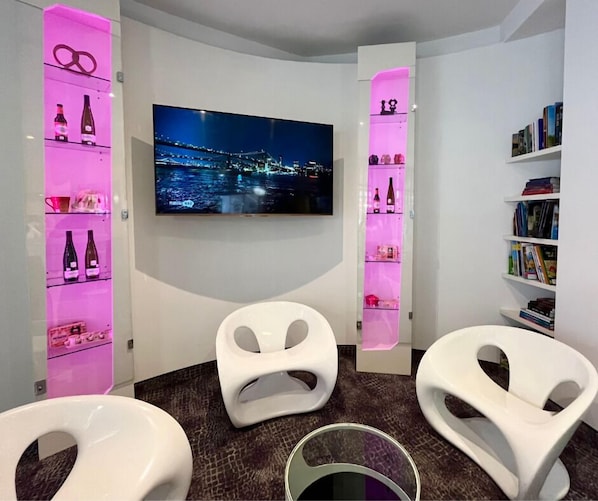 A modern room with a flat-screen TV displaying a cityscape, two white chairs, a glass table, and shelves with books and bottles.