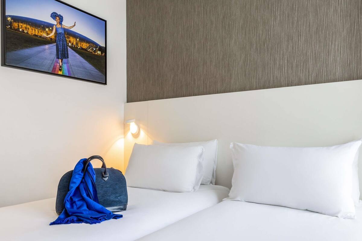 A hotel room with a bed, white pillows, a blue bag, and a wall-mounted picture.
