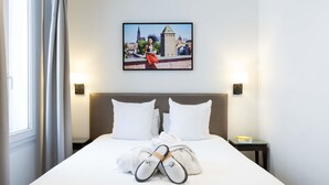 Une chambre d’hôtel avec un grand lit, deux oreillers, un tableau encadré au mur et une paire de chaussons sur le lit.