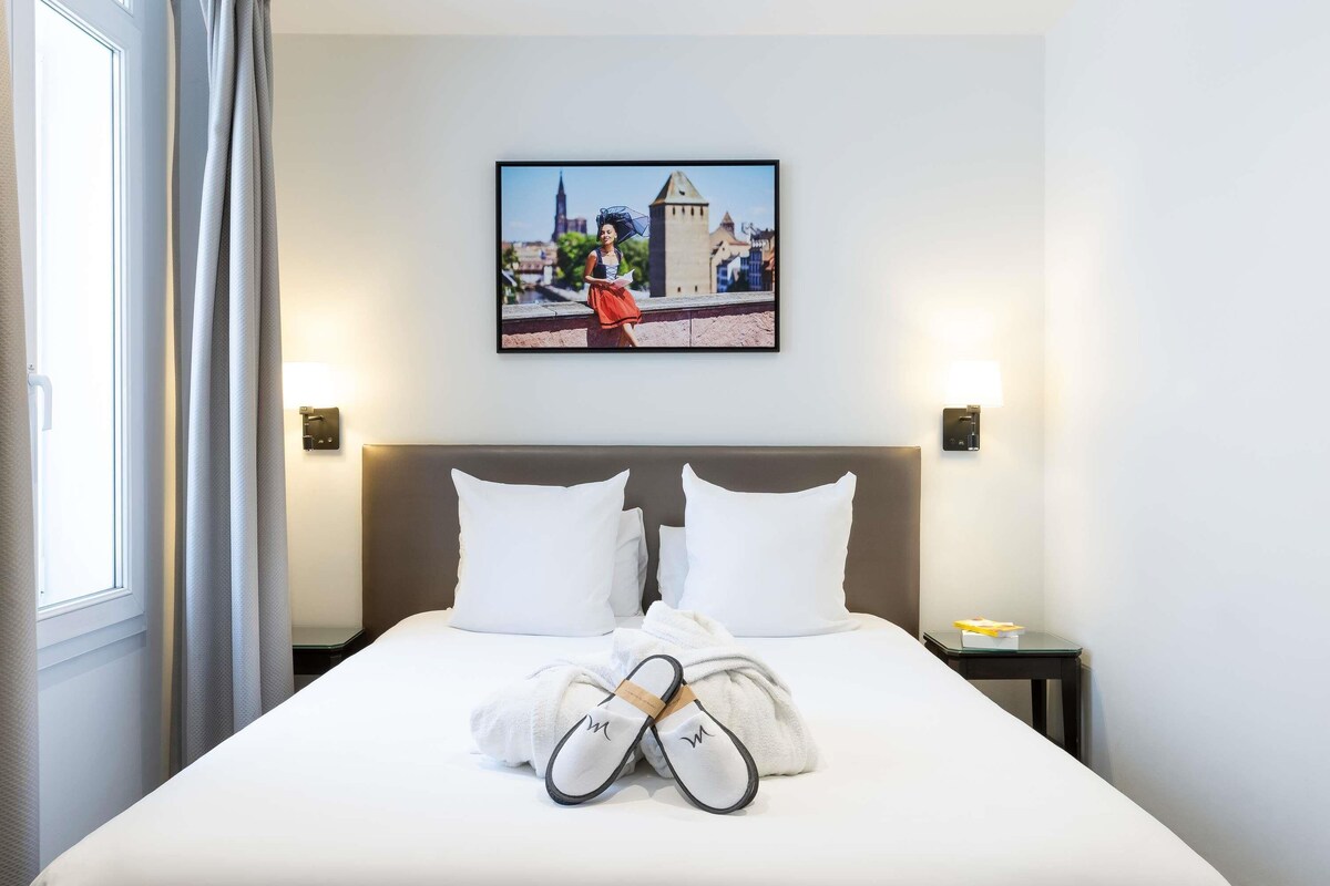 A hotel room with a large bed, two pillows, a framed picture on the wall, and a pair of slippers on the bed.