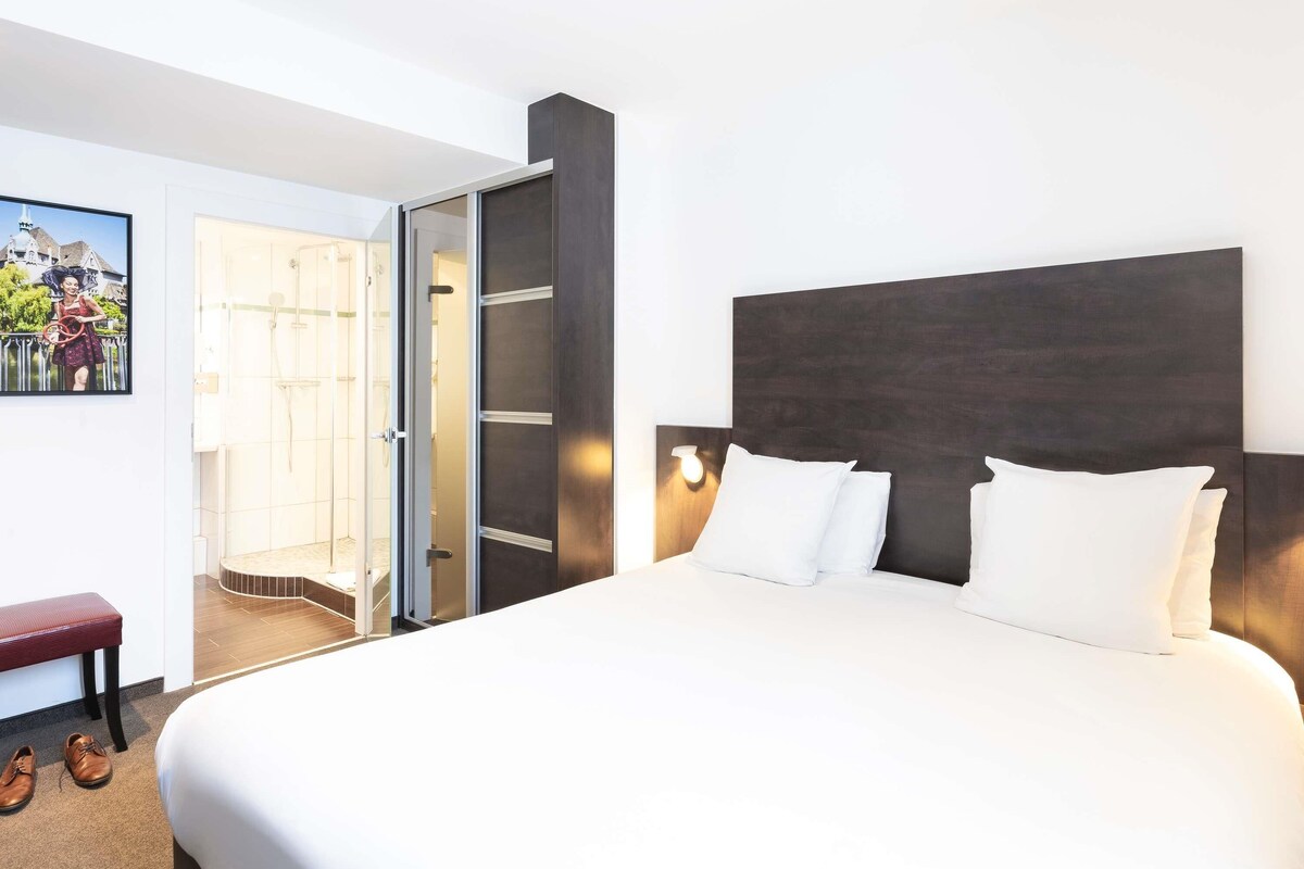Une chambre d’hôtel avec un grand lit, un banc, une paire de chaussures, une porte donnant sur une salle de bain et un luminaire fixé au mur.