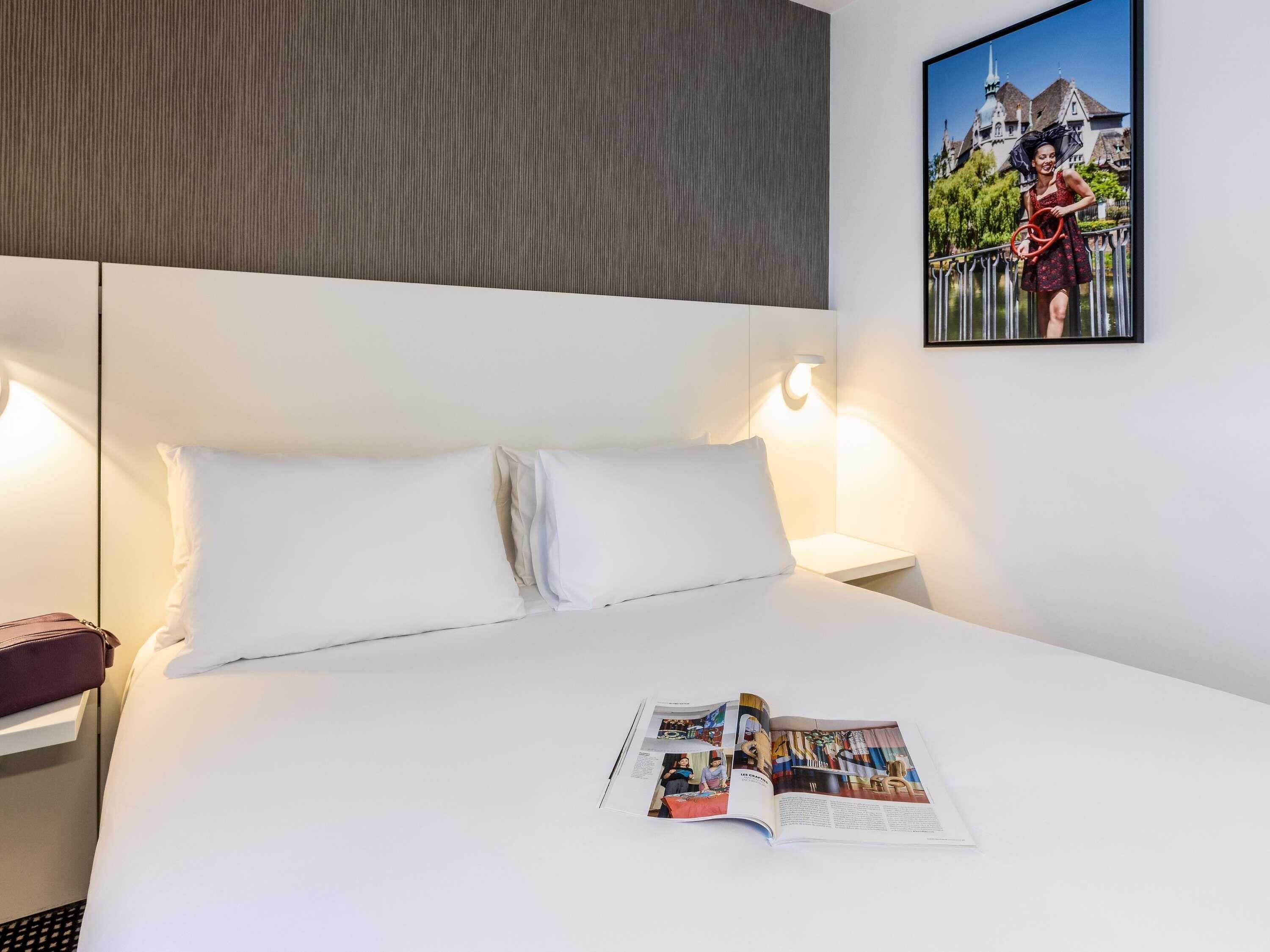 A hotel room with a bed, white bedding, a magazine on the bed, a nightstand, a lamp, and a framed picture on the wall.