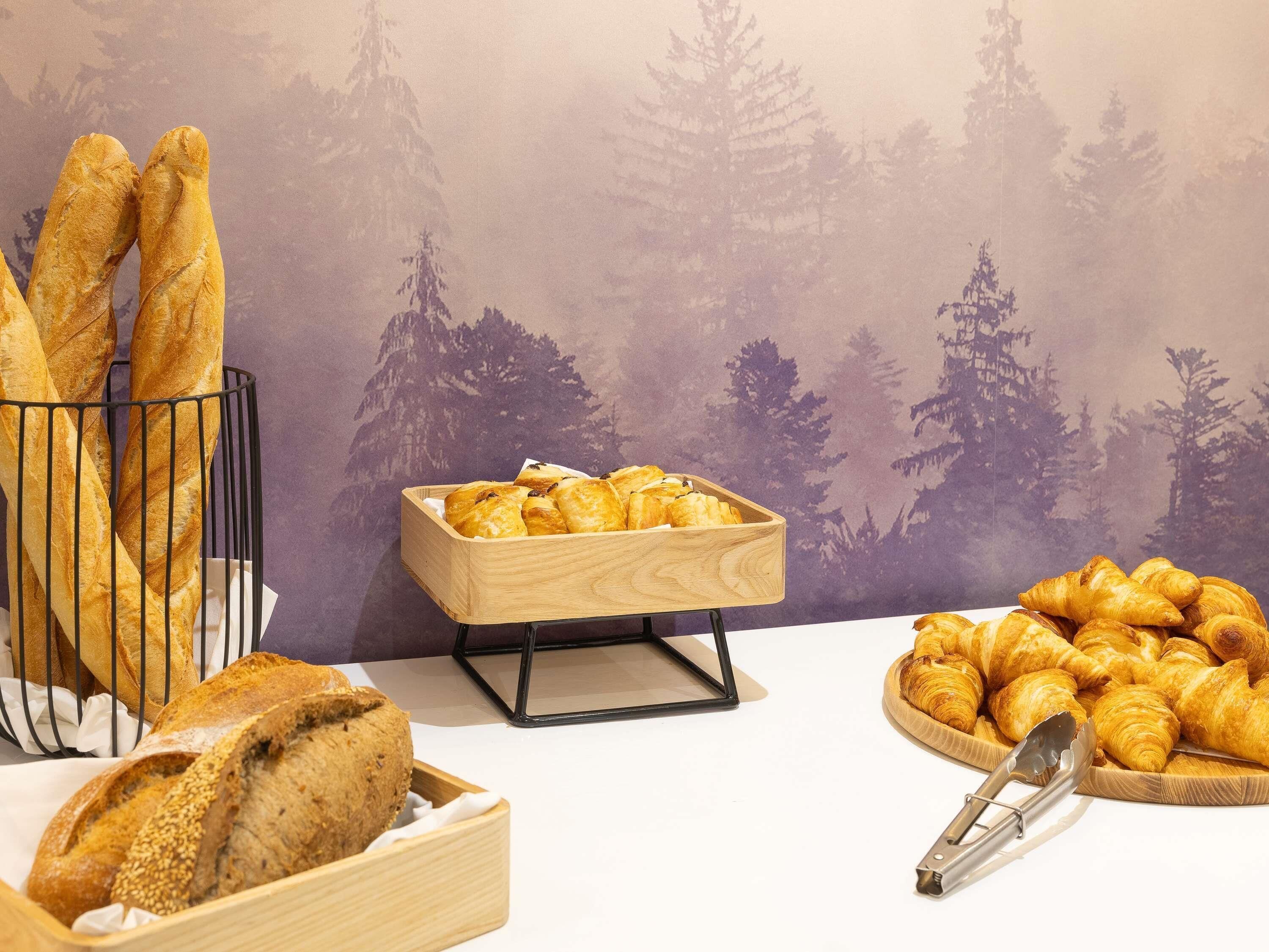 A variety of breads and pastries displayed in wooden containers and a metal rack, with a forest-themed backdrop.