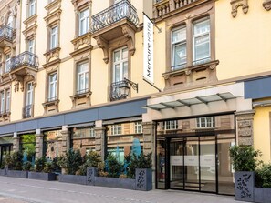 Entrada de un hotel con puerta de cristal, un letrero sobre la puerta y un balcón con barandillas de hierro forjado.