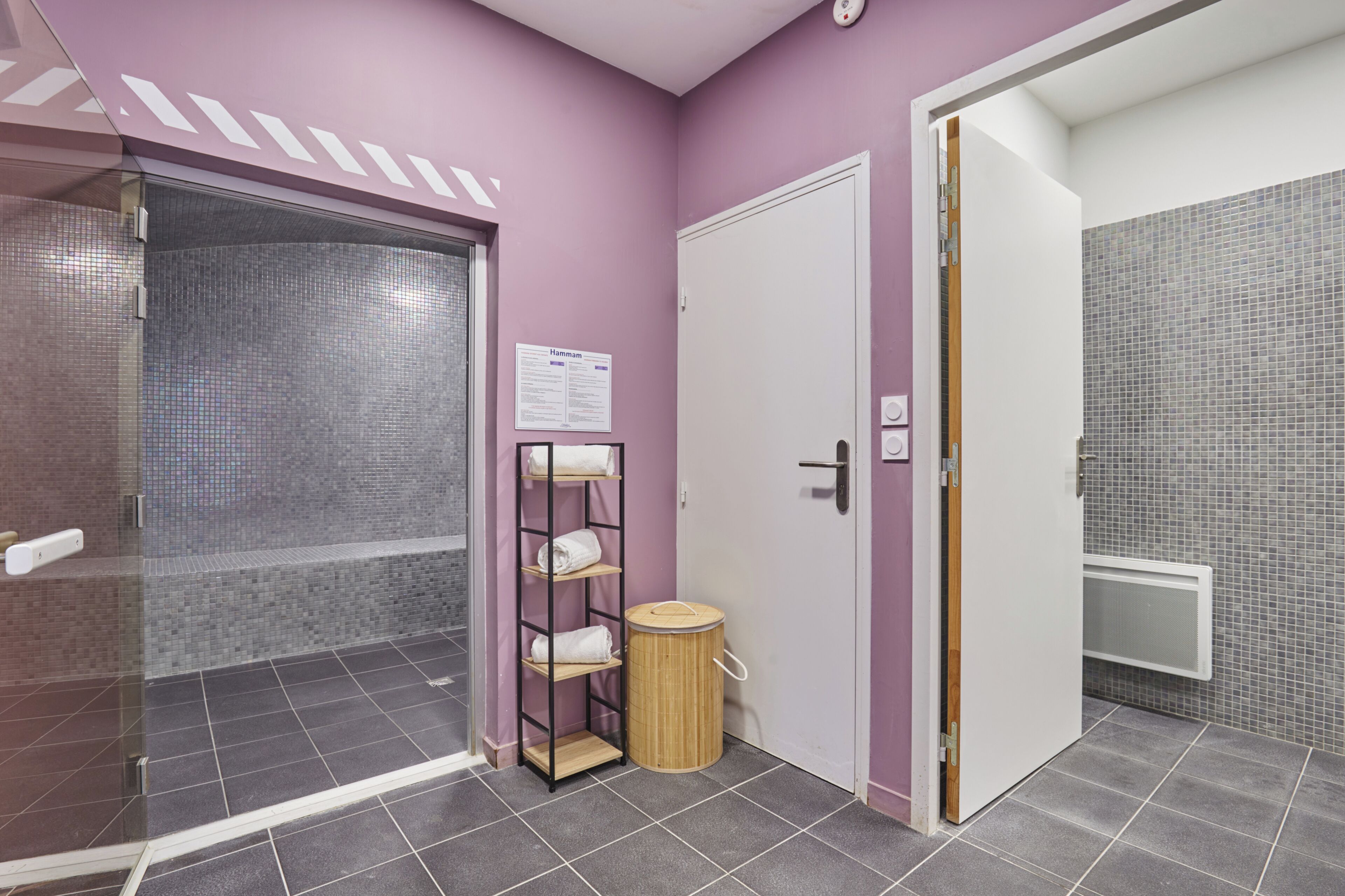 A bathroom with a tiled floor, a shower area, a white door, and a shelf with towels.