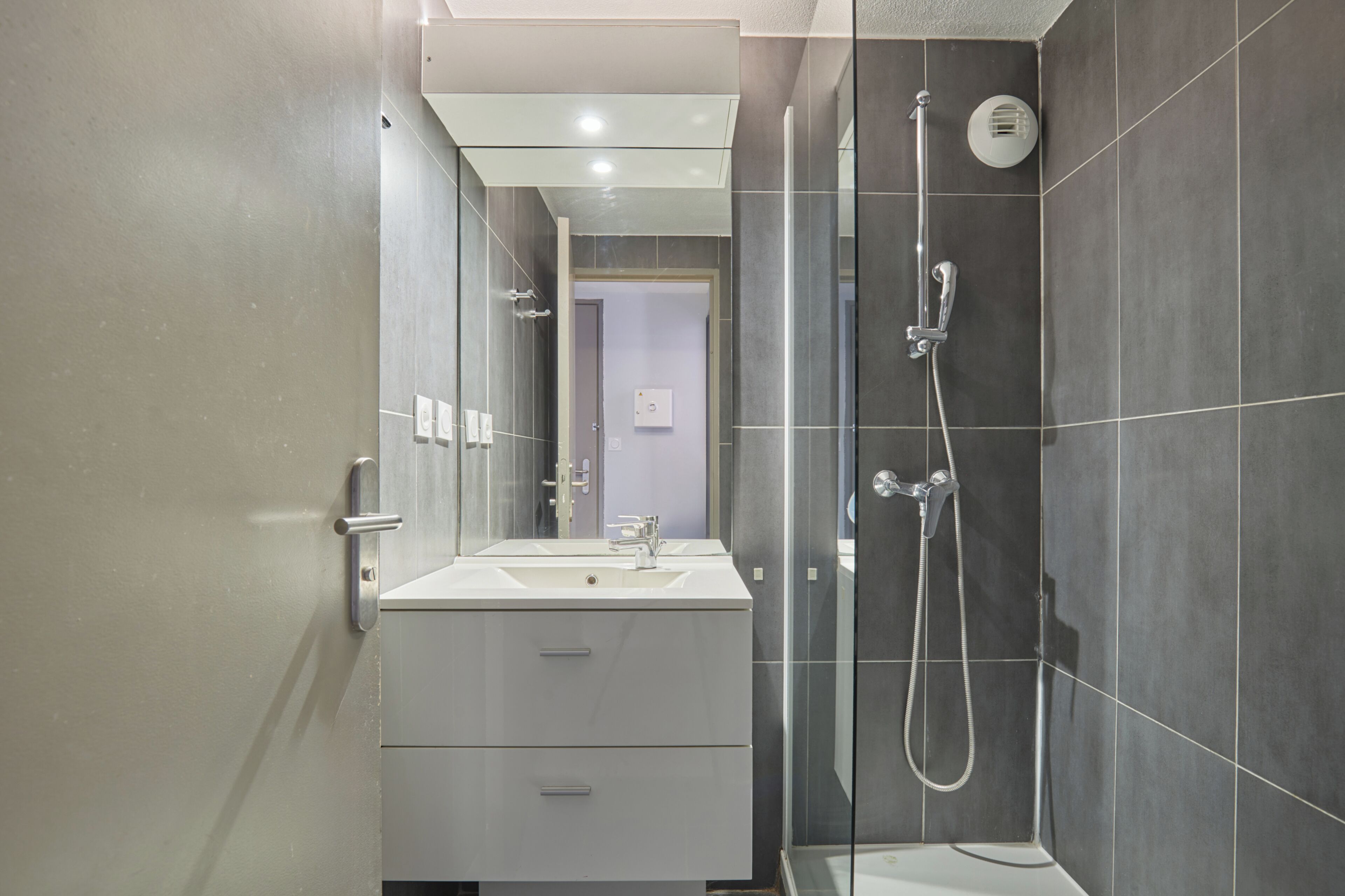 A modern bathroom with a white sink, a glass shower enclosure, and dark tiled walls.