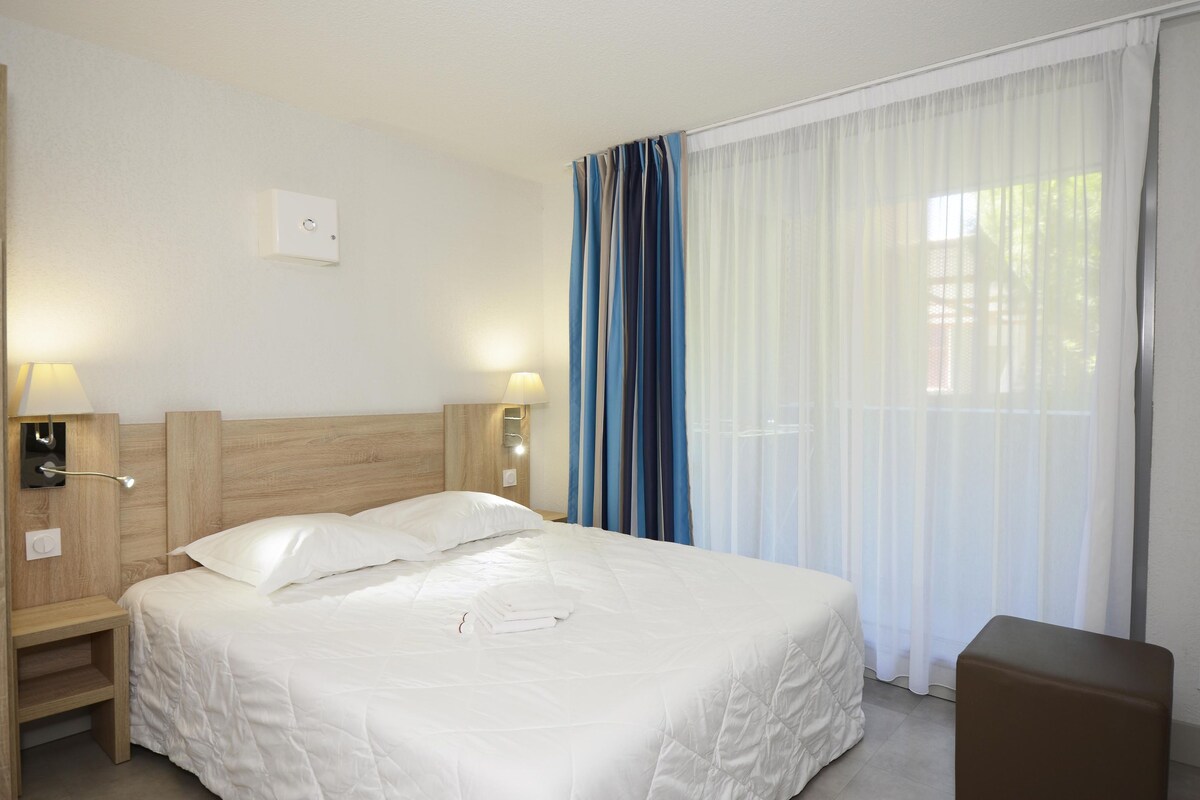 A hotel room with a bed, bedside lamps, a nightstand, a wooden headboard, a window with sheer curtains, and a small brown ottoman.
