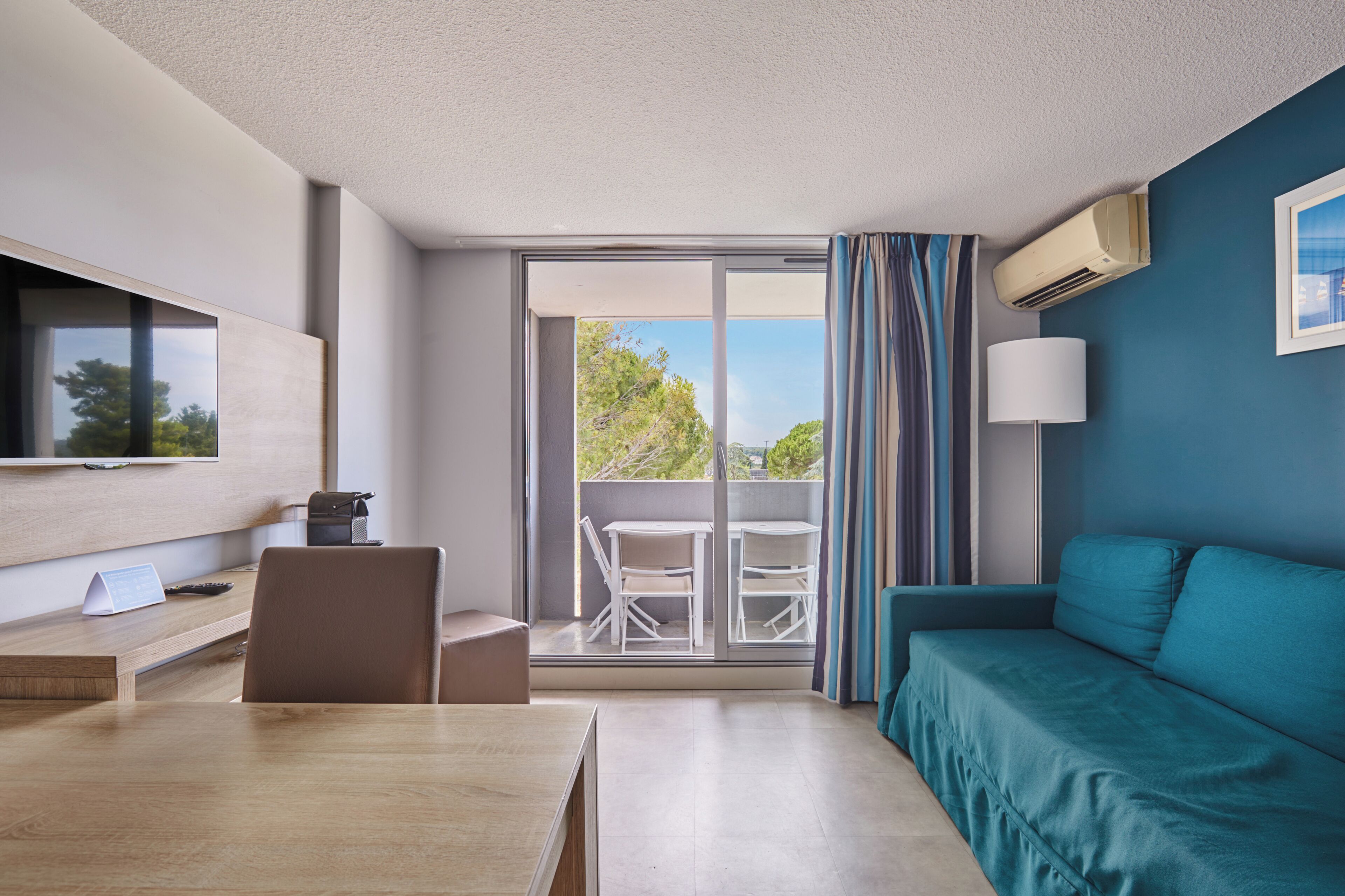 A hotel room with a balcony, a TV, a desk, a sofa, and a lamp.