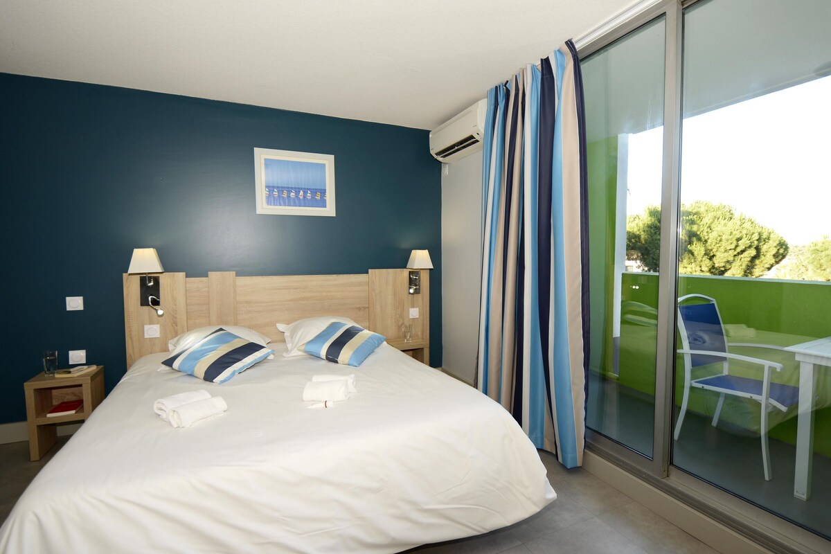 A hotel room with a bed, two bedside lamps, a nightstand, a window with curtains, and a view of greenery outside.