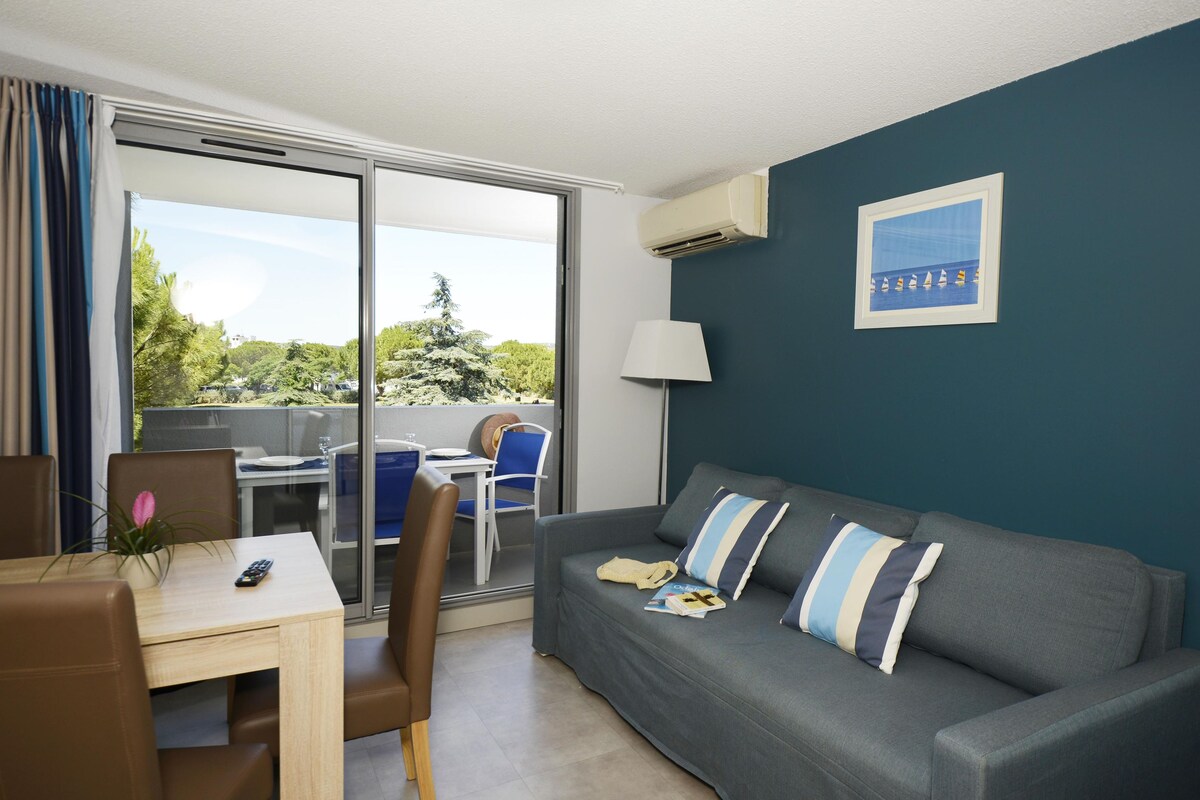 A modern living room with a grey sofa, a dining table, and a chair. There is a view of a balcony with a glass door, and a painting on the wall.