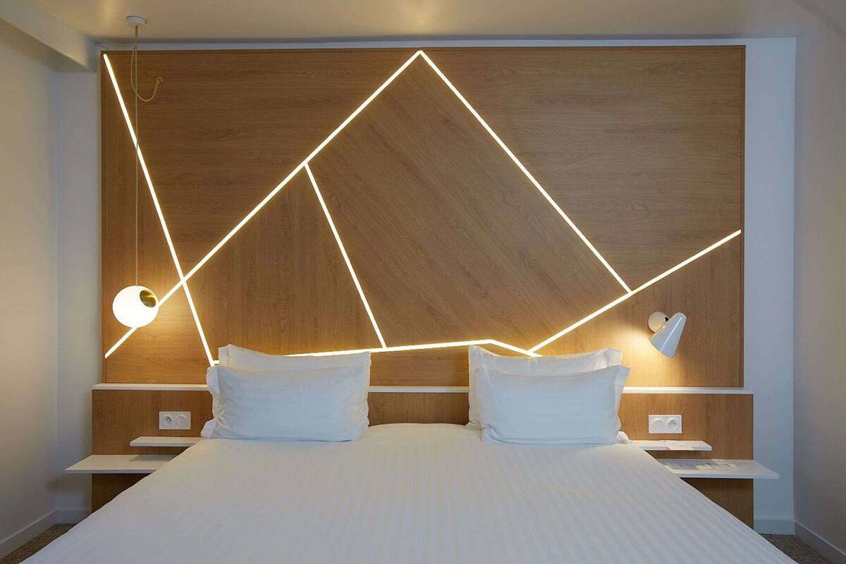 A modern hotel room with a large bed, wooden headboard, bedside tables, and a wall-mounted light fixture.