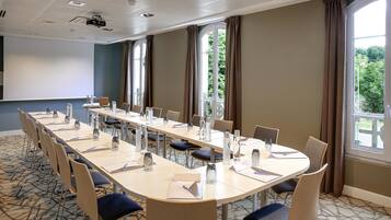 A conference room with a long table, chairs, a projector screen, and large windows with curtains.