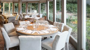 Un restaurante con mesas redondas preparadas para comer, ventanas amplias que dan a zonas verdes y una decoración de estilo moderno.