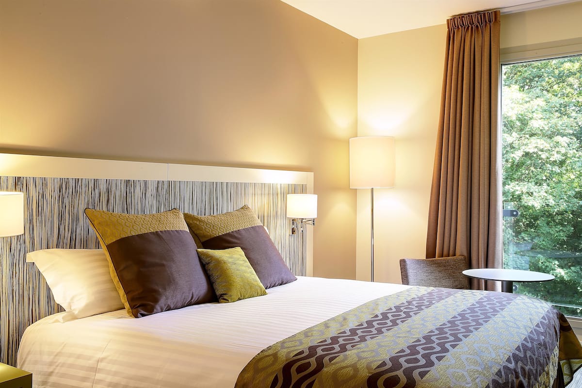 A hotel room with a large bed, two bedside lamps, a window with curtains, and a view of greenery outside.