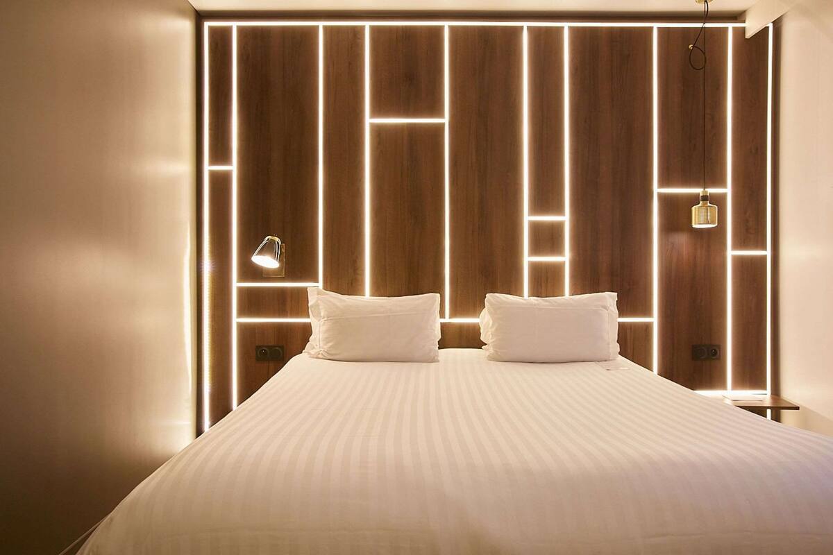 A modern hotel room with a large bed, a bedside lamp, and a wooden headboard with illuminated panels.