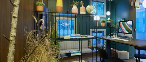 A modern office space with a desk, computer, and books. There are potted plants and a large window.