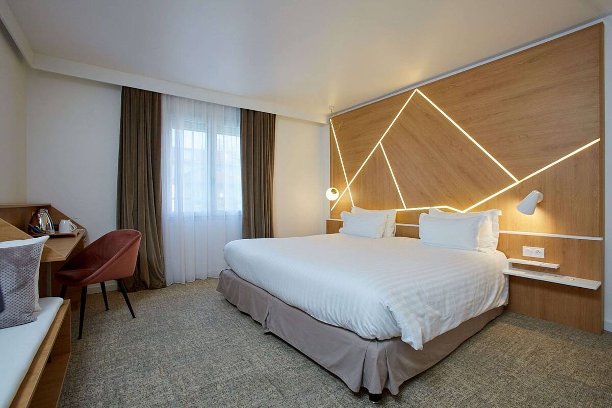 A hotel room with a large bed, a desk with a chair, a nightstand, and a wall-mounted lamp.
