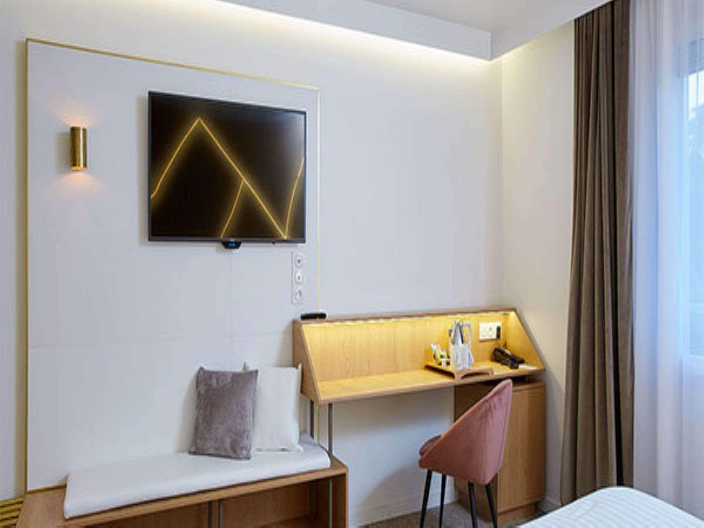 A modern hotel room with a bed, a desk with a chair, a TV mounted on the wall, and a window with curtains.