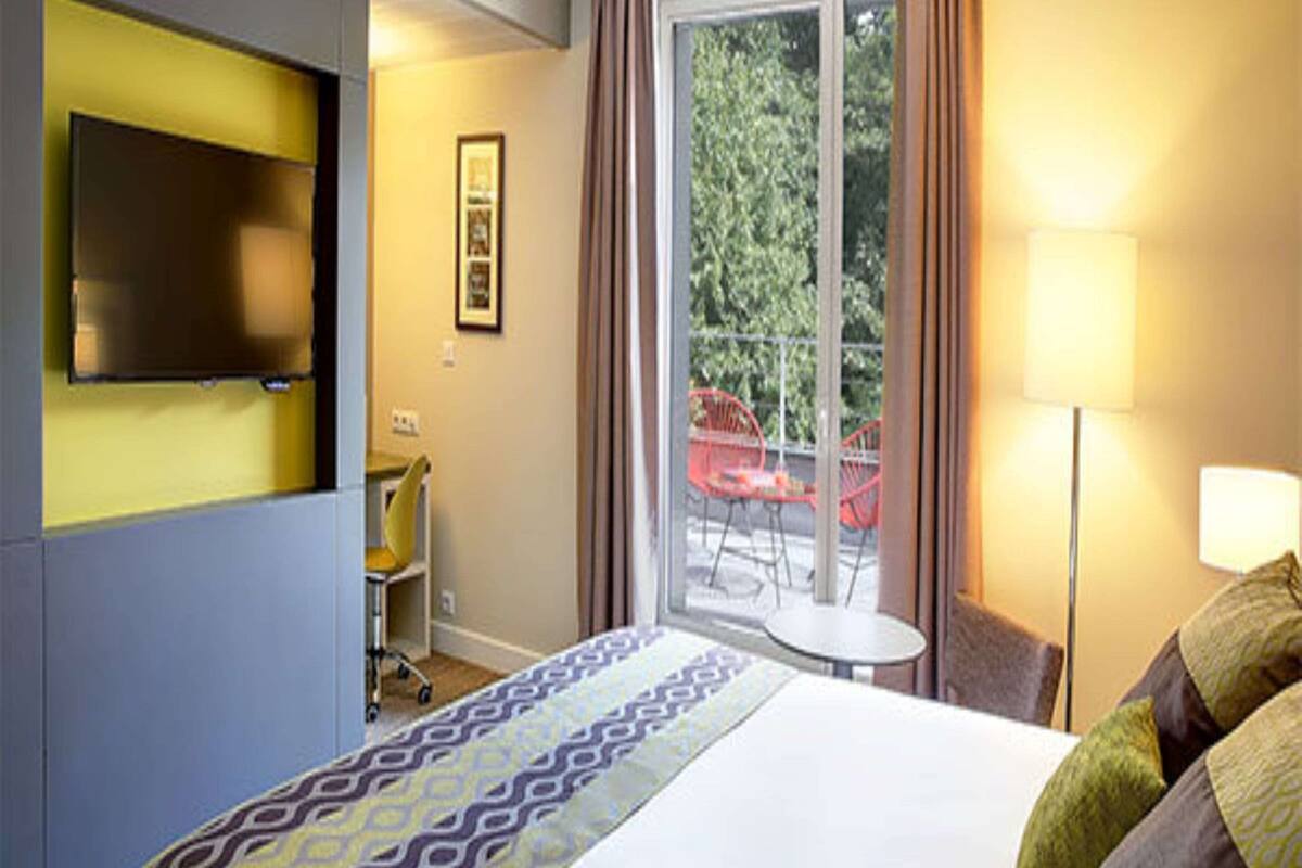 A hotel room with a bed, a desk, a chair, a TV, a window with curtains, and a balcony with chairs.