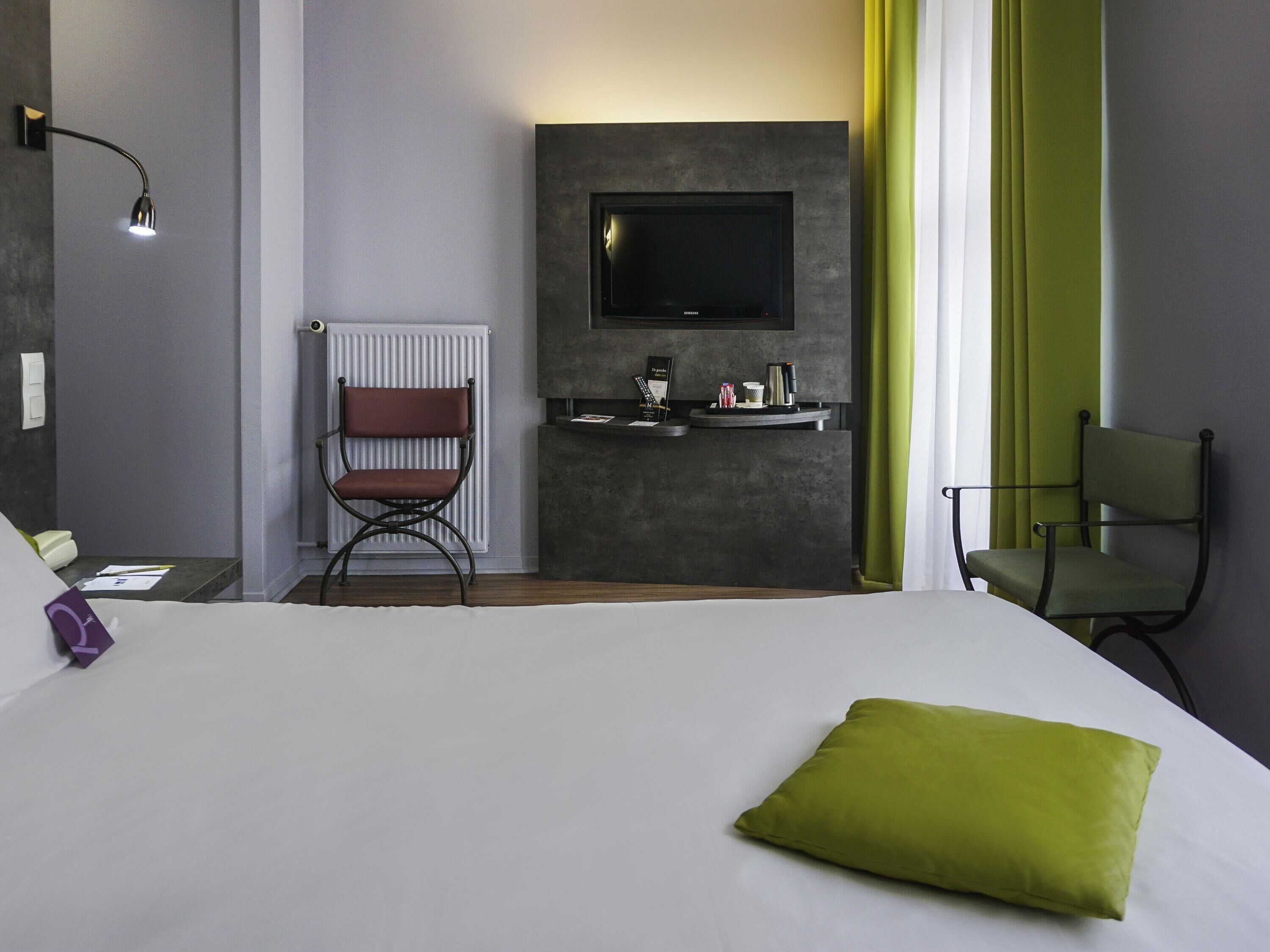 A hotel room with a bed, a chair, a desk, a television, and a radiator.
