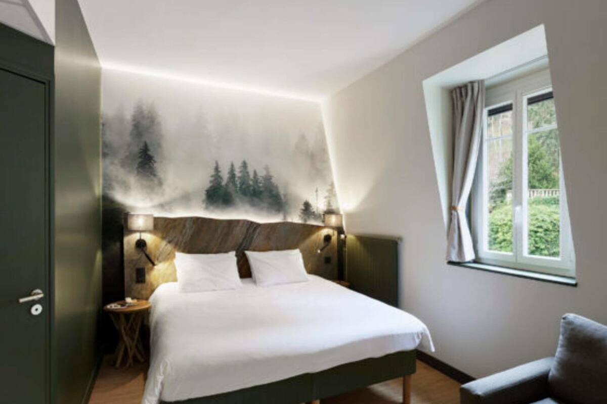 A bedroom with a bed, nightstands, a window, and a view of greenery.
