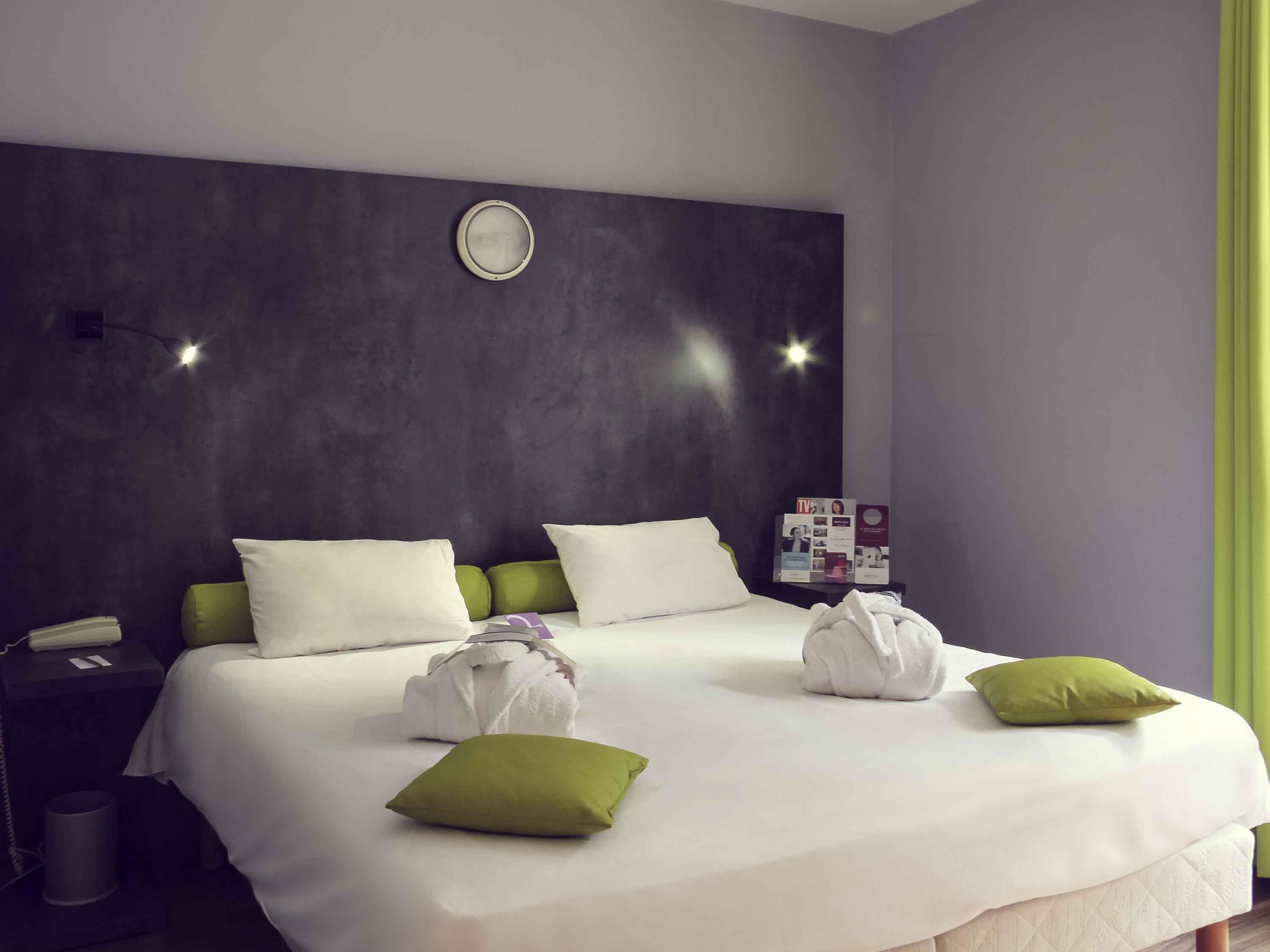 A hotel room with a large bed, two pillows, a bedside table with a phone, a trash bin, and a wall clock.