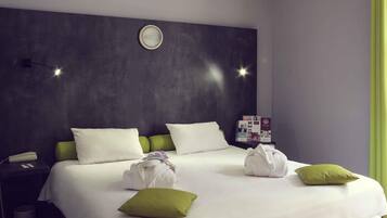 A hotel room with a large bed, two pillows, a bedside table with a phone, a trash bin, and a wall clock.