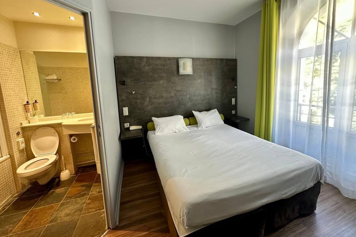 A modern hotel room with a large bed, a bedside table, a nightstand, a bathroom with a toilet, sink, and mirror.
