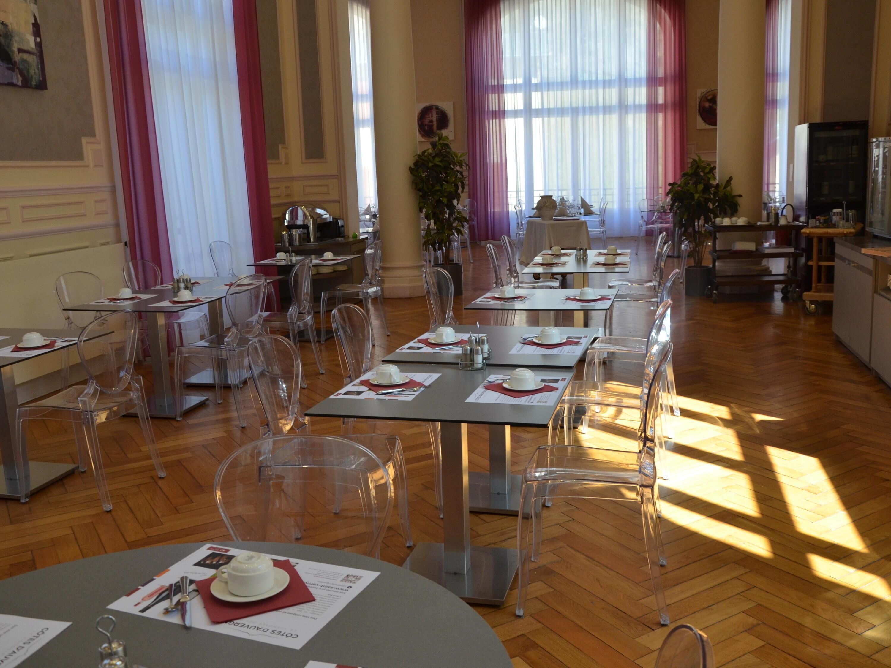 A dining area with clear chairs and tables, wooden floors, and large windows with curtains.