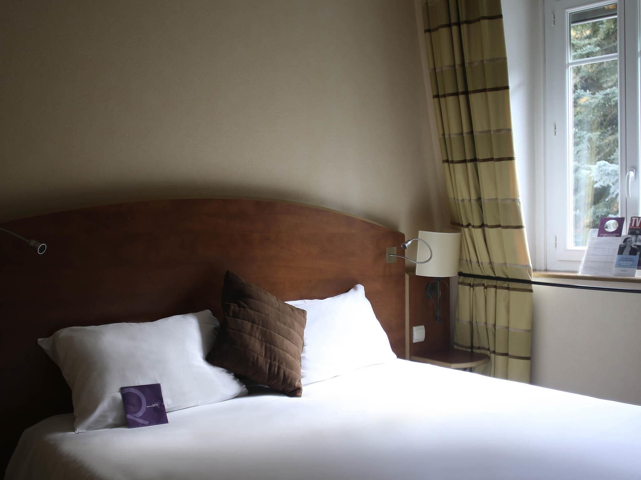 Habitación de hotel con cama, cabecero, mesita de noche, lámpara, ventana con cortinas y vistas a árboles.