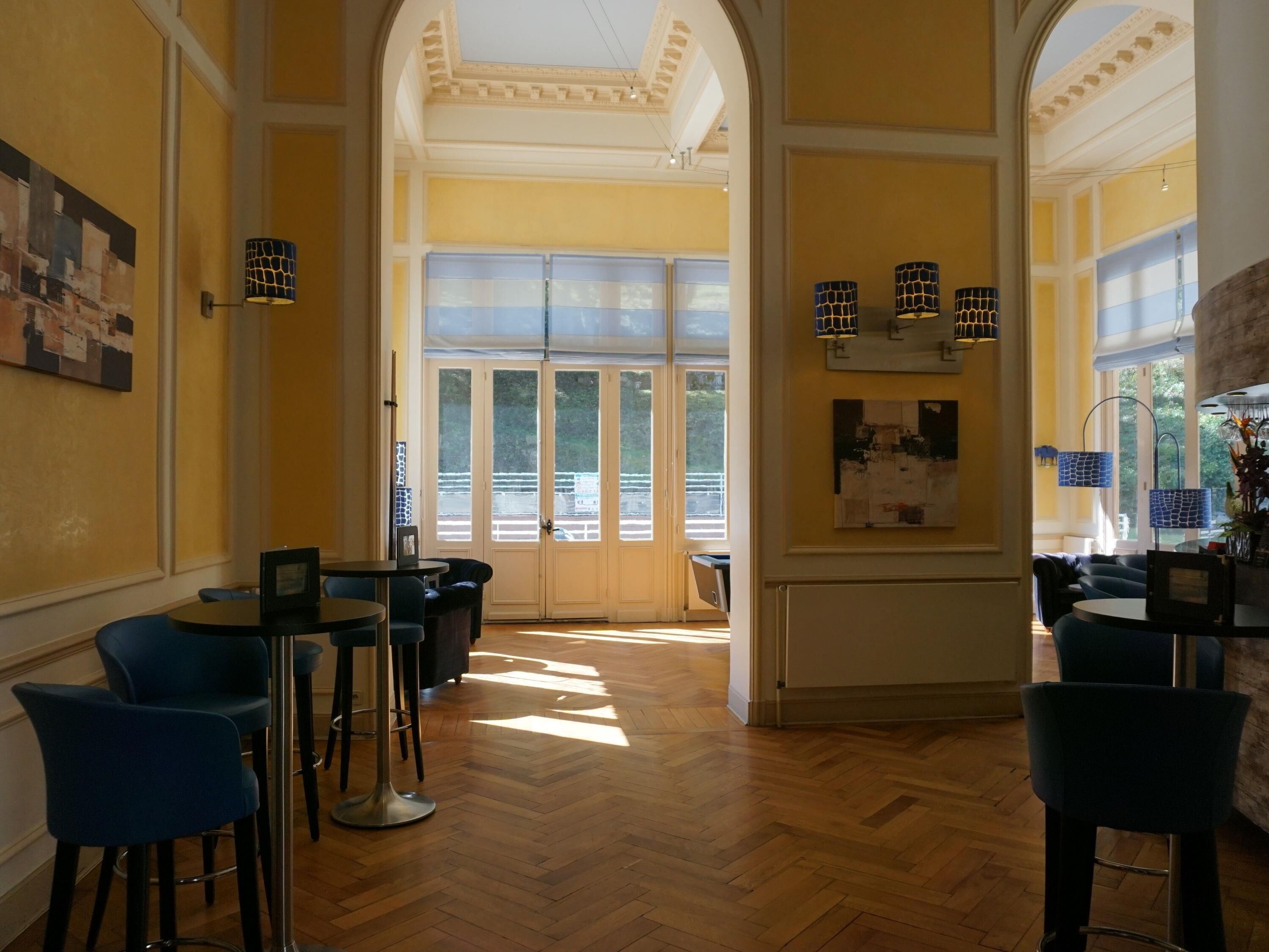 Un hall d’hôtel doté d’un parquet en bois à motifs chevrons, de chaises bleues, d’un coin bar et de grandes fenêtres laissant entrer la lumière naturelle.