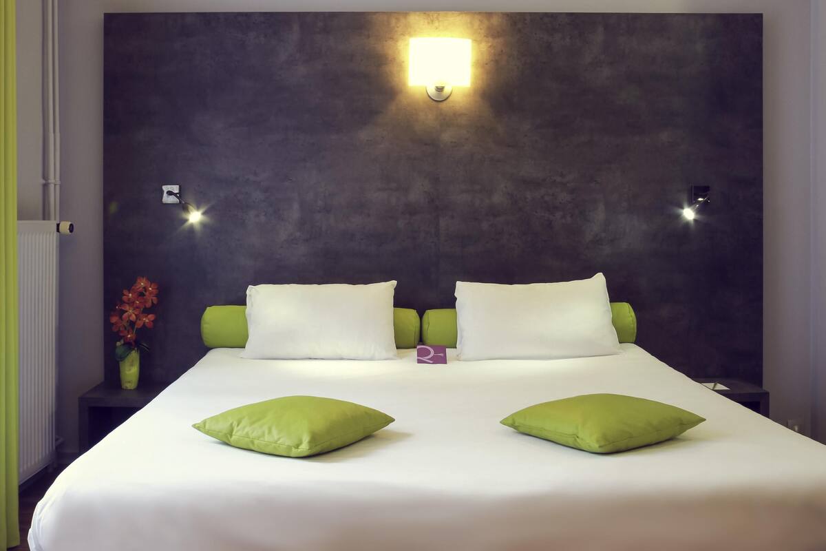 A hotel room with a large bed, two pillows, a nightstand, a vase with flowers, and a wall-mounted light fixture.