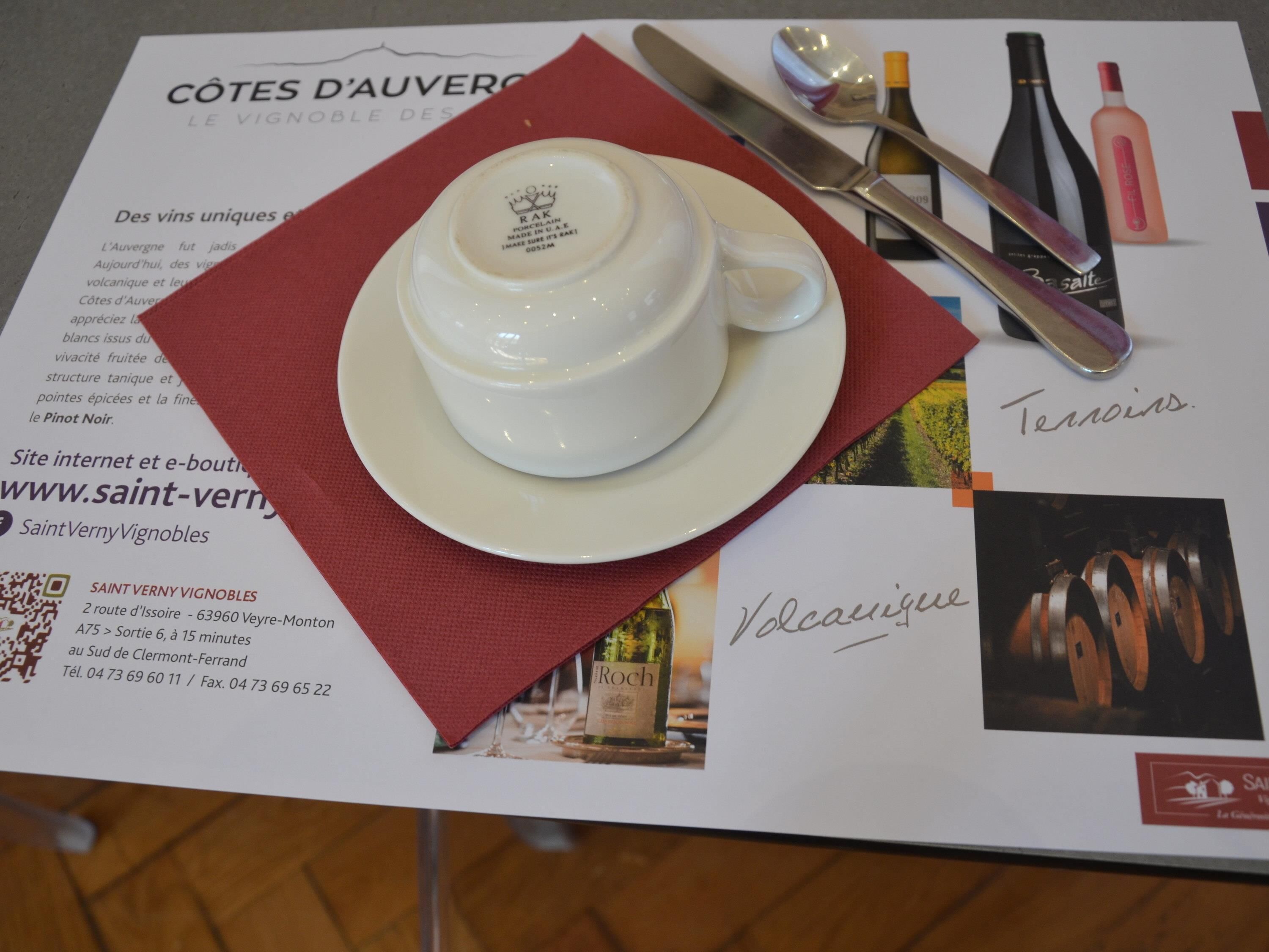 Una mesa con una tetera blanca en un platito, un mantel rojo y un menú con imágenes y texto sobre botellas de vino.