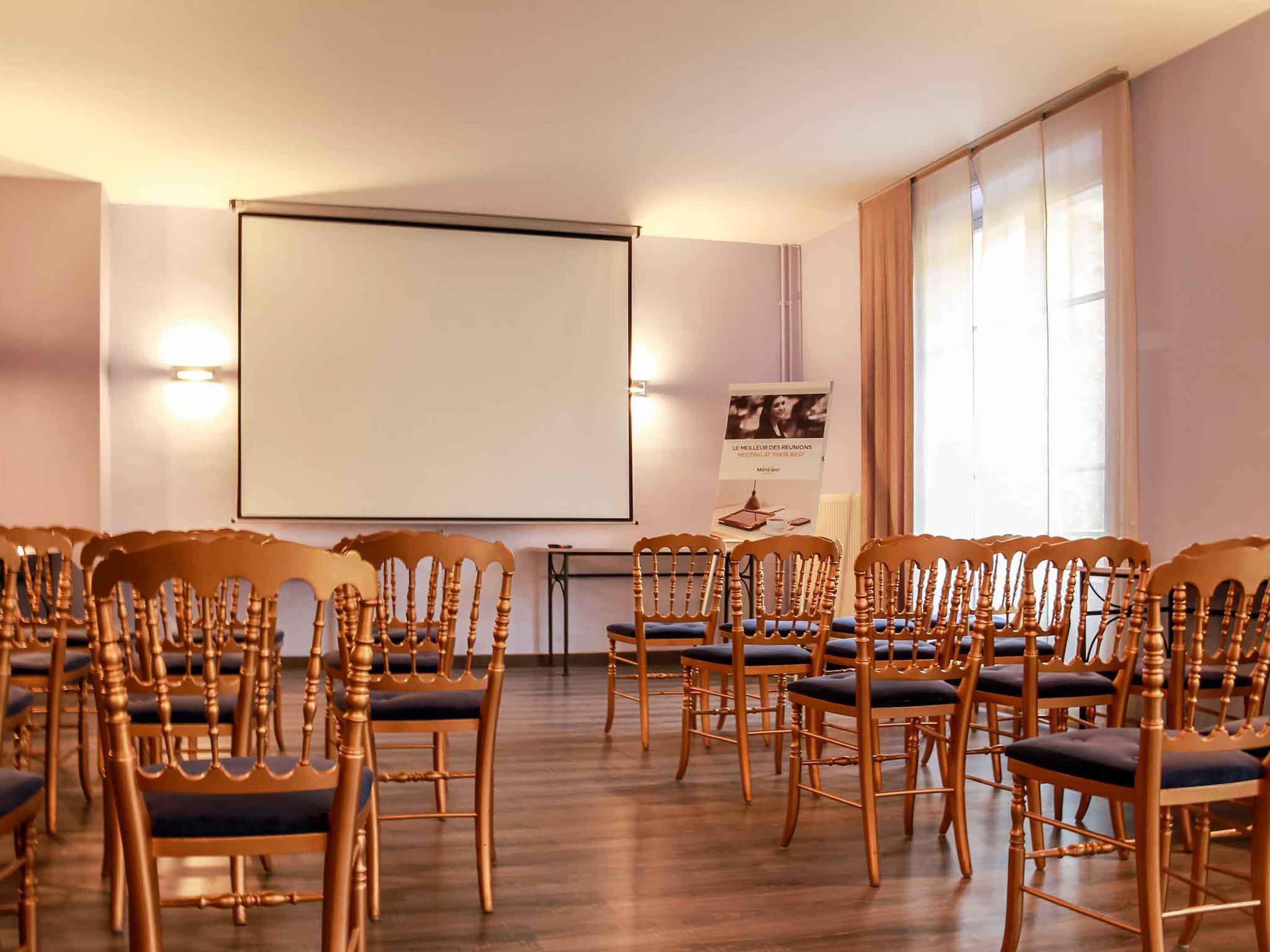 A conference room with rows of wooden chairs, a projector screen, and a poster on the wall.