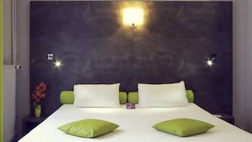A hotel room with a large bed, green pillows, a nightstand with a vase of flowers, and a wall-mounted light fixture.