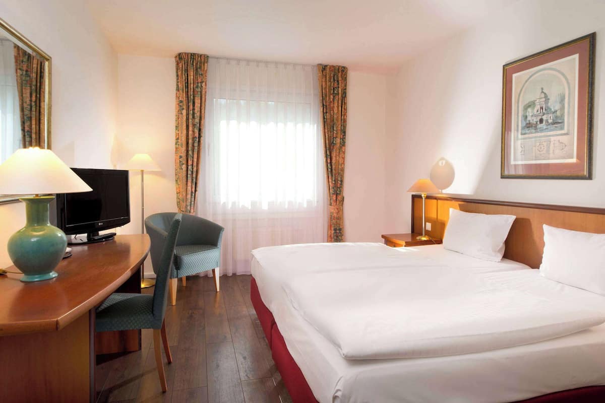 Une chambre d’hôtel avec un lit, un bureau avec une lampe, une chaise, un téléviseur et un tableau encadré au mur.