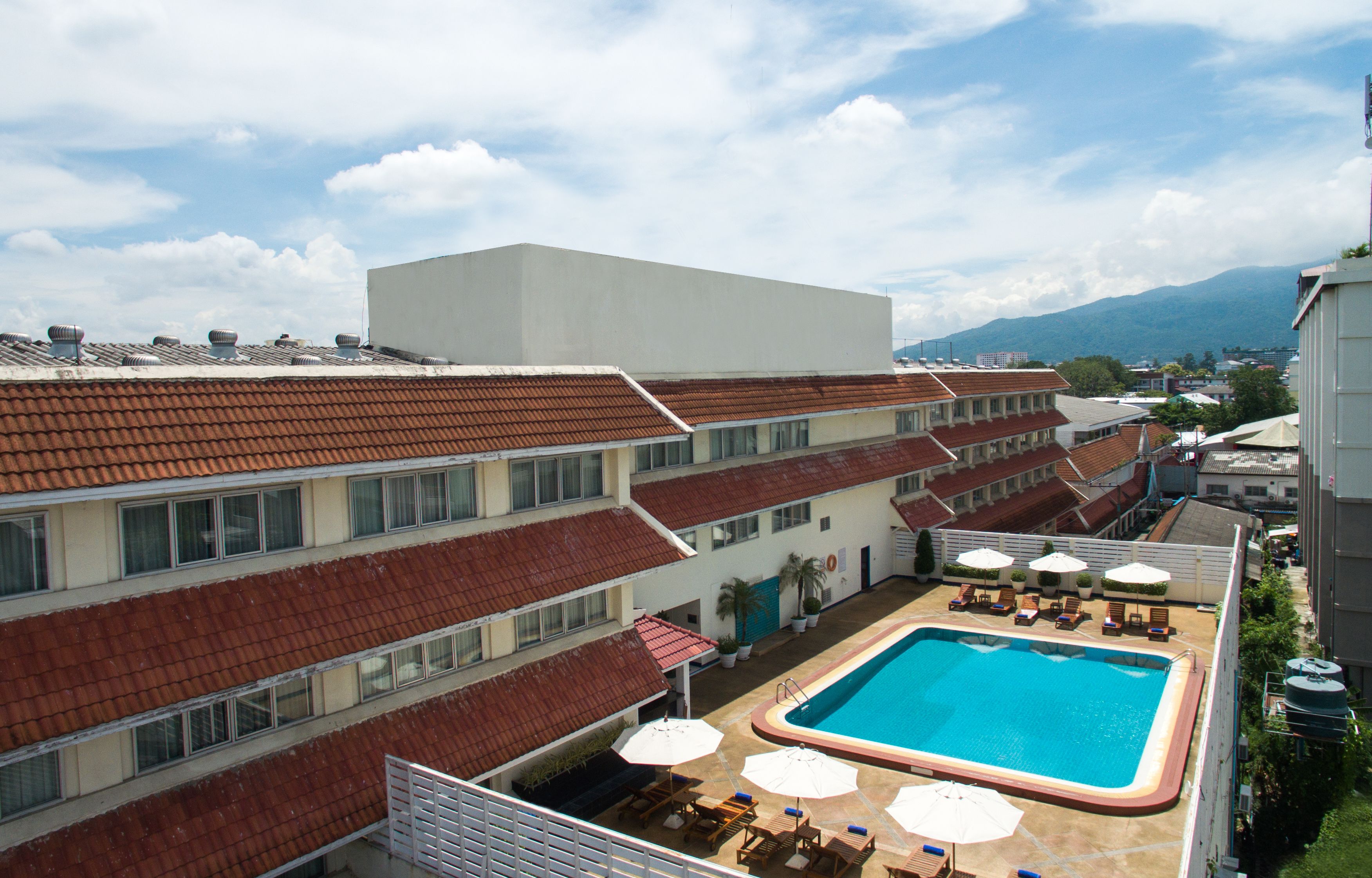 Una piscina en la azotea con sillones y sombrillas, rodeada de edificios con techos de tejas rojas.