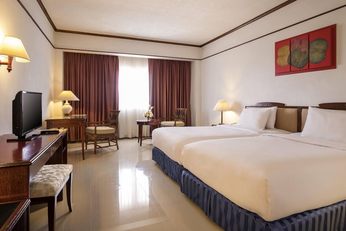 A hotel room with a large bed, a desk with a television, a chair, a lamp, and a window with curtains.