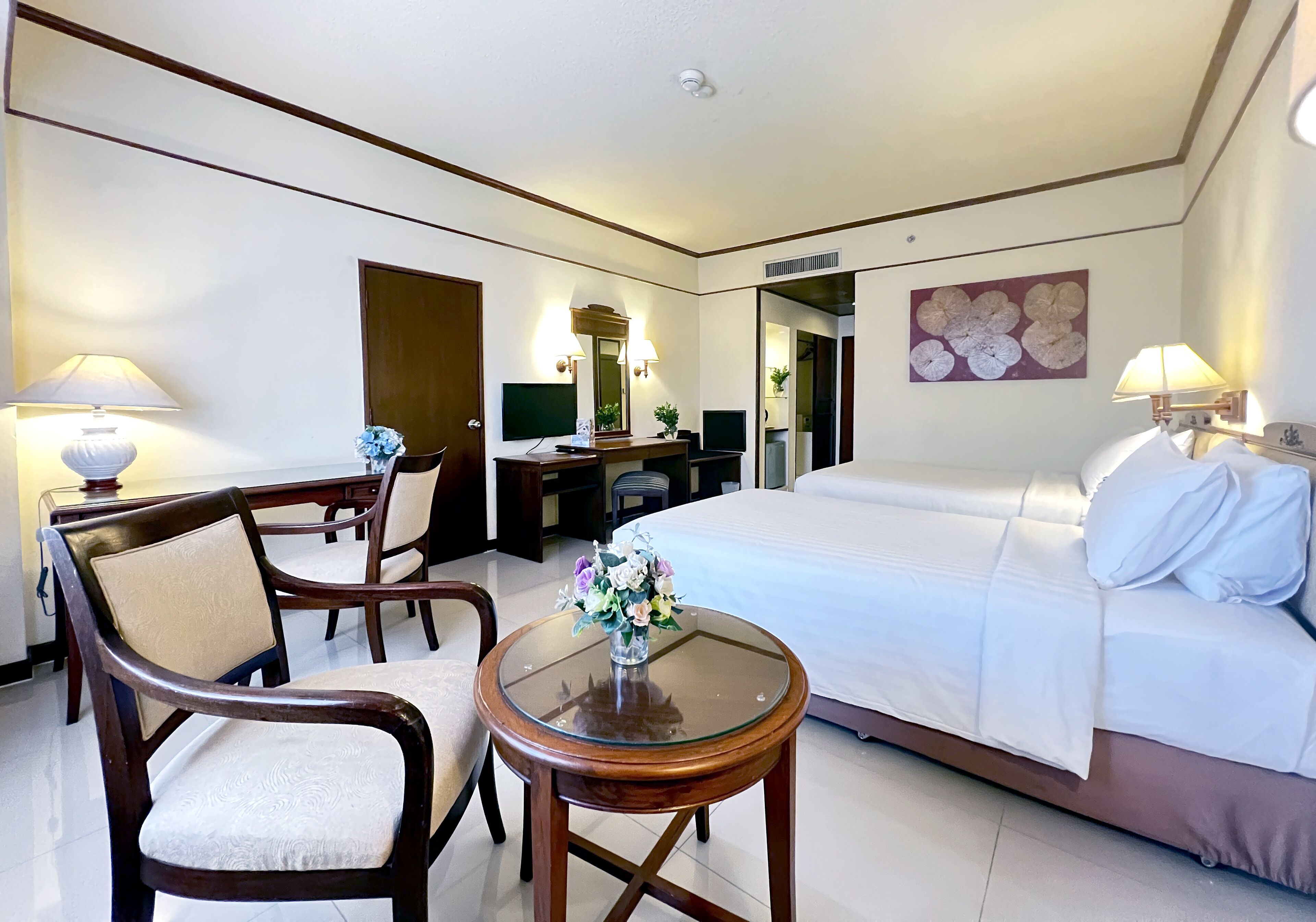 A hotel room with a bed, a desk, two chairs, a small table with a vase of flowers, and a television.