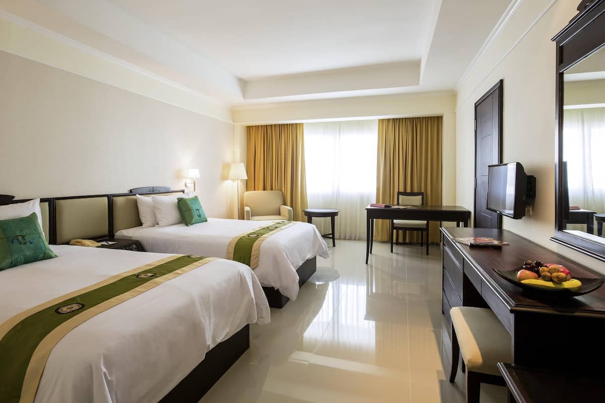 A hotel room with two beds, a desk, a chair, a television, and a dining table with fruit.