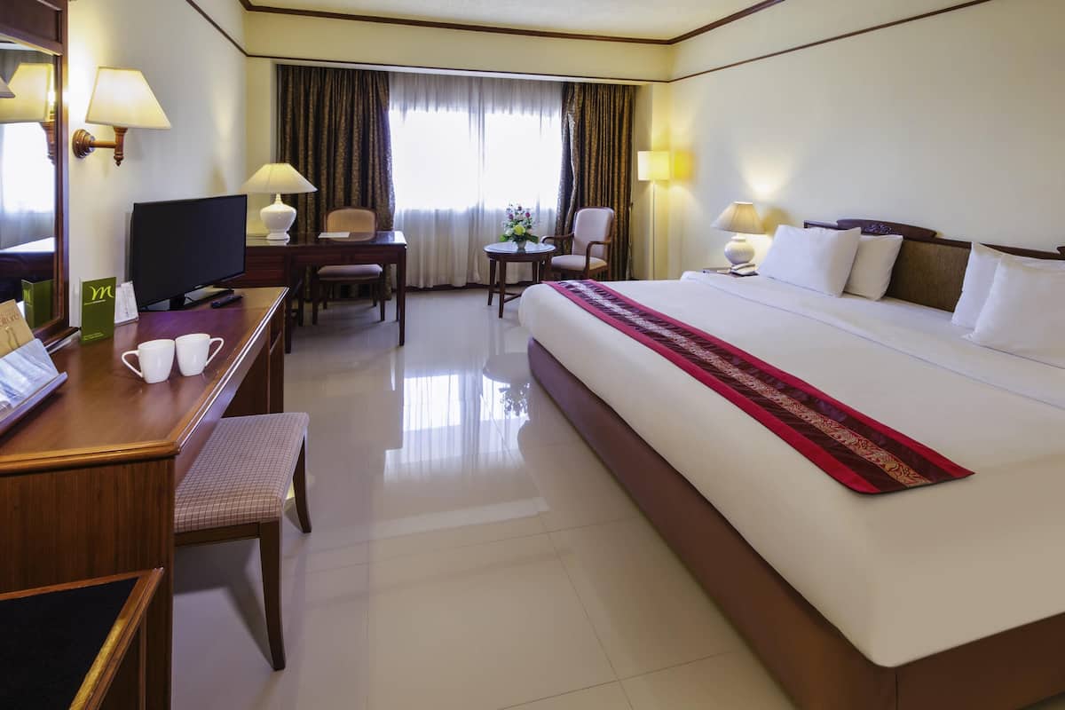 A hotel room with a large bed, a desk with a computer, a chair, a small table with a lamp, and a window with curtains.