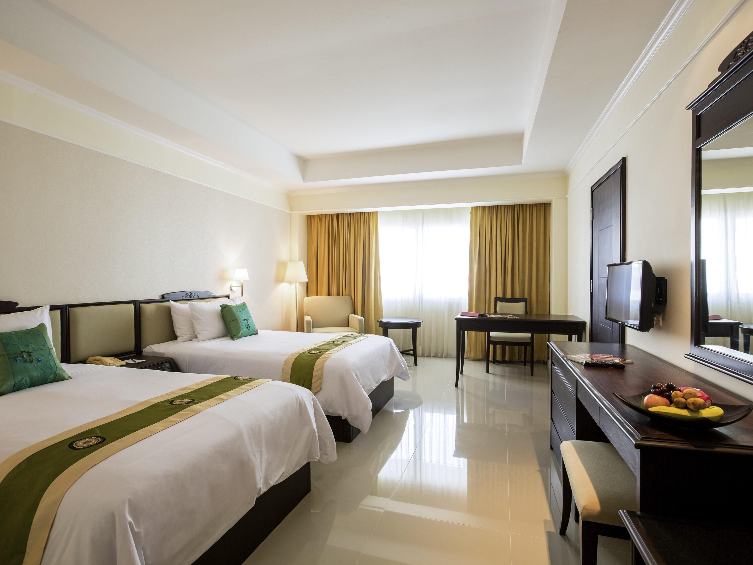 A hotel room with two beds, a desk, a chair, a television, and a dining table with fruit.
