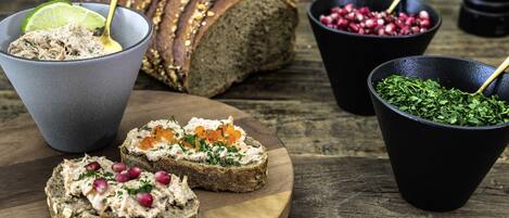 A wooden board with two slices of bread topped with a creamy spread and pomegranate seeds, a loaf of bread, and two small black bowls containing herbs and pomegranate seeds.