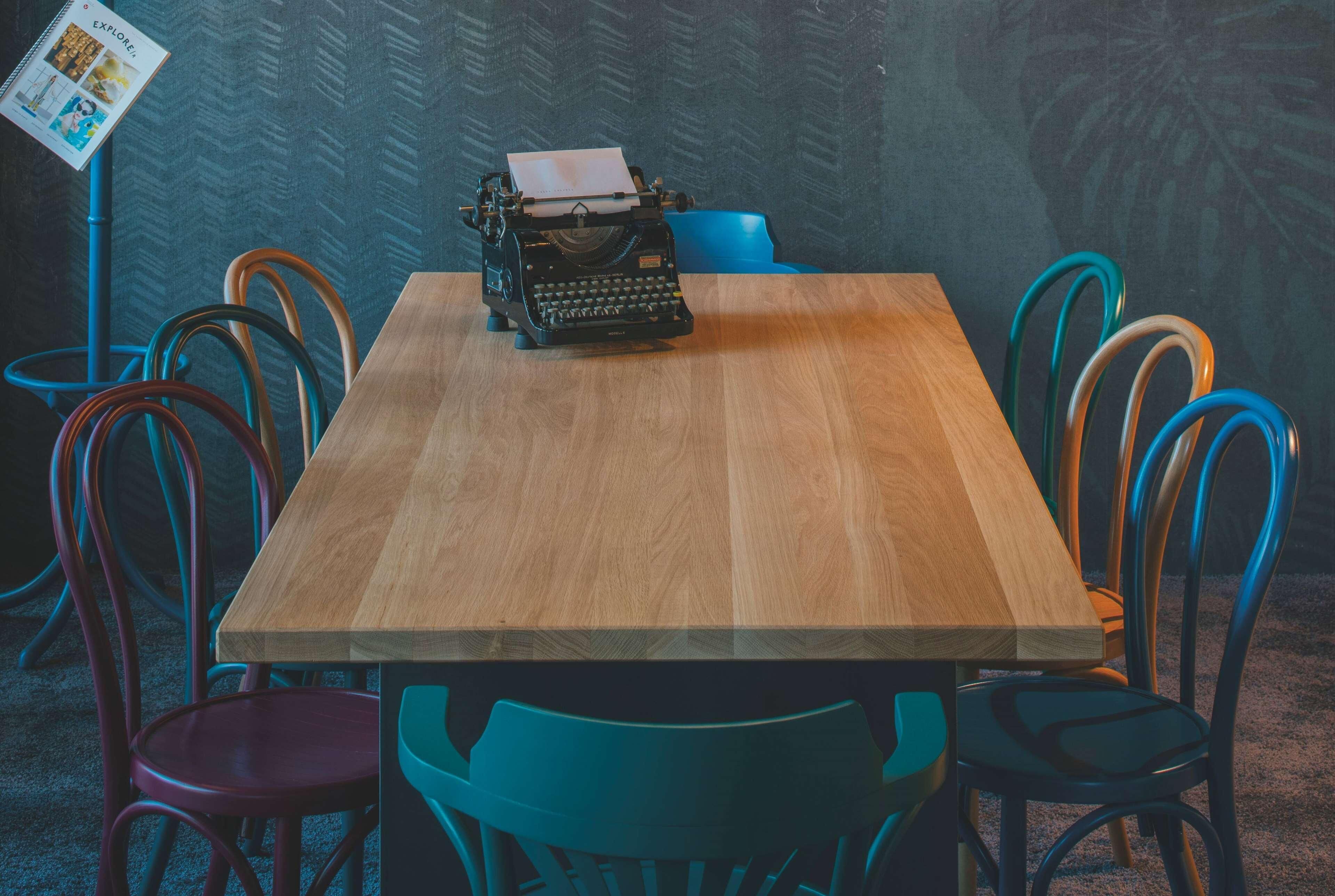 A wooden table with a typewriter, surrounded by colorful chairs.
