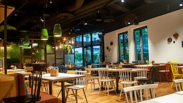Ein Restaurant mit Holztischen und -stühlen, hängenden grünen Pendelleuchten und großen Fenstern mit Blick auf viel Grün.
