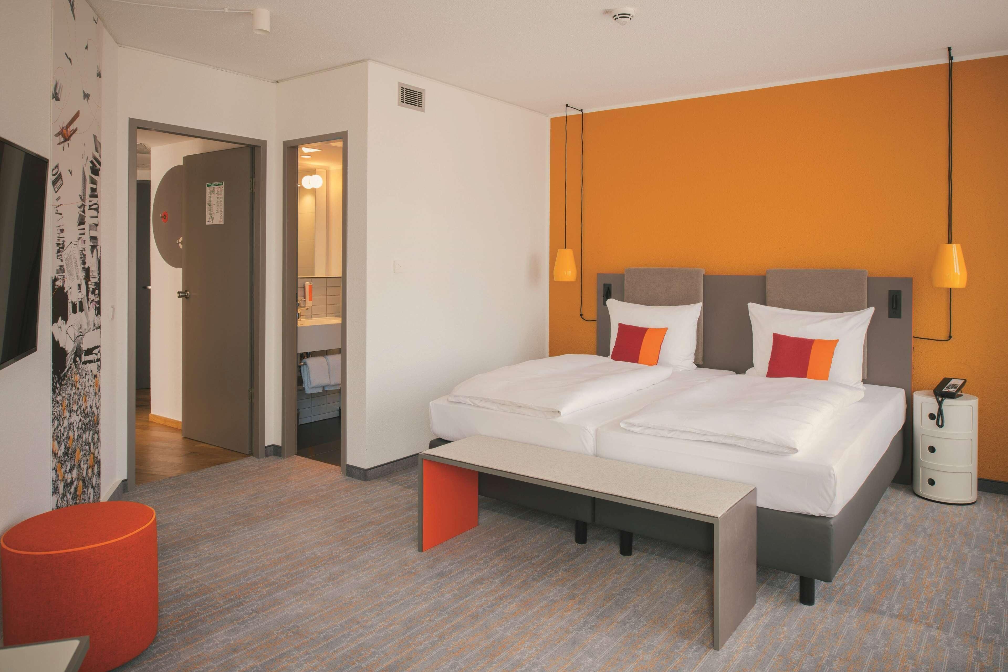 A modern hotel room with a double bed, a bedside table, a red ottoman, a nightstand with a phone, and a bathroom visible through an open door.